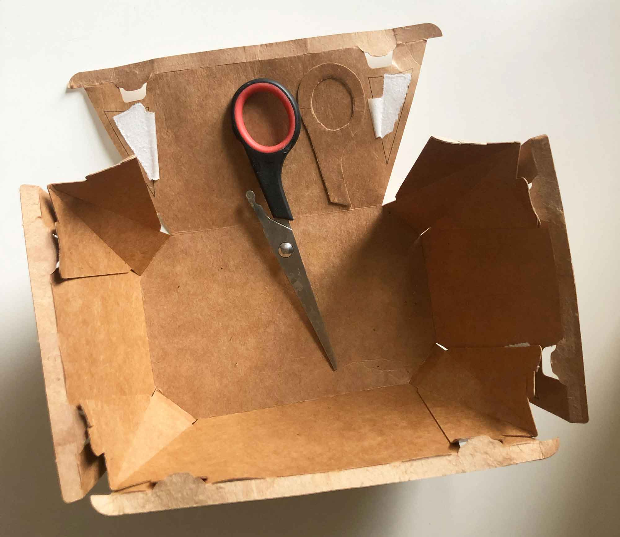 photograph of a flatten cardboard food carton with scissors with one broken handle and a cardboard template of the handle 